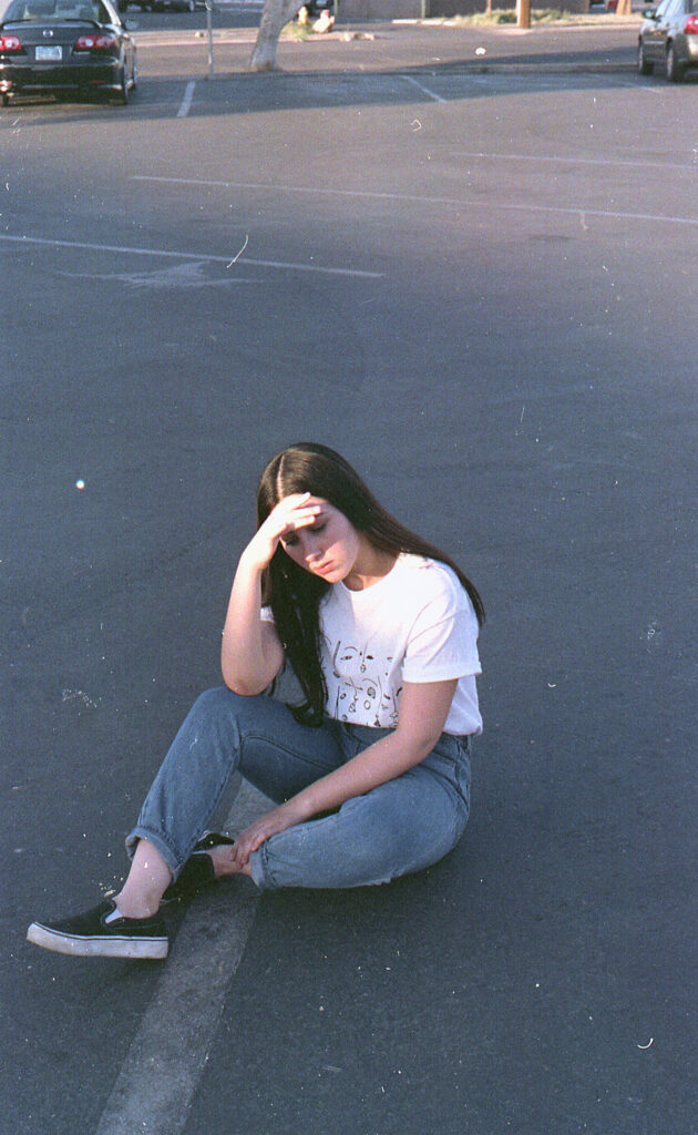 Film Photo of Gabriela Inzunza sitting in a parking lot wearing a white t-shirt with a graphic design blue jeans and black sneakers. She's holding her forehead with one hand looking down and there are parked cars in the background.